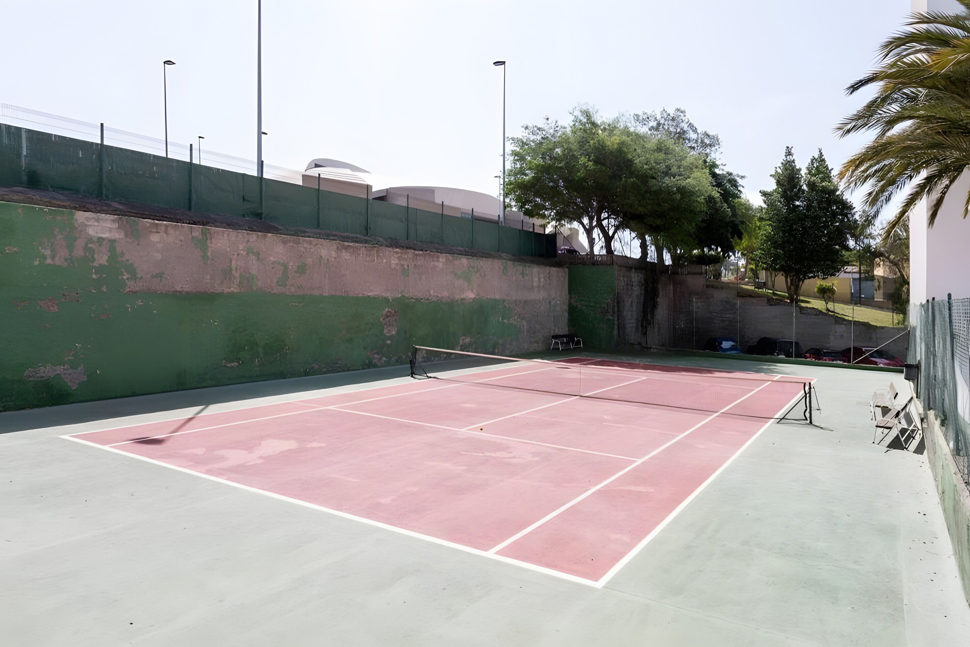 Een tennisbaan buiten, met een groene muur en bomen op de achtergrond.