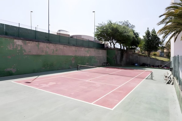 Een tennisbaan buiten, met een groene muur en bomen op de achtergrond.