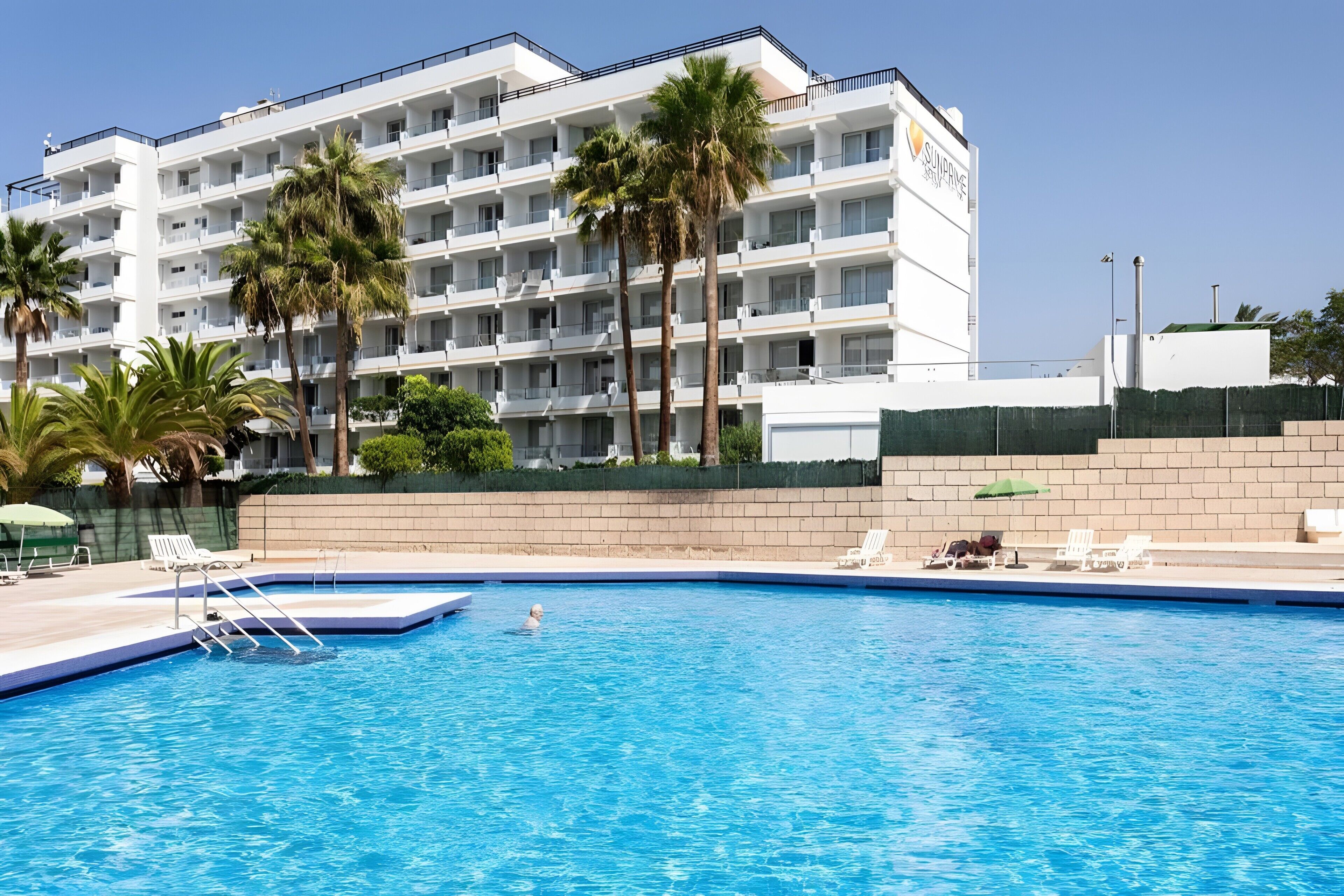A swimming pool with a clear blue water surface, surrounded by lounge chairs and a white building with multiple balconies.