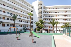 A playground with swings and a slide, surrounded by palm trees and a multi-story building with balconies. - Vacation Apartment 'Cozy  Playa Las Americas' with Sea View, Balcony and Wi-Fi (Costa Adeje)