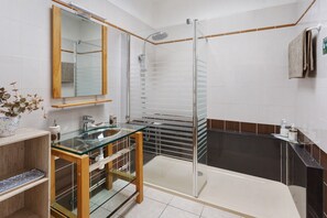 A modern bathroom with a glass-enclosed shower, a wooden vanity with a glass top, and a large mirror. - Chambre Iris - Demeure Olea (Chérac)