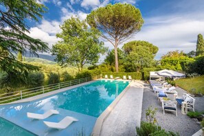 A swimming pool with a clear blue water surface, surrounded by a white fence. There are lounge chairs and umbrellas arranged for relaxation. - Refined And Luxurious Villa In the Outskirts Of Lucca (Lucca)
