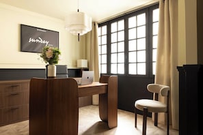 Une chambre d’hôtel avec un bureau en bois, un ordinateur portable, un vase avec des fleurs, une chaise et une grande fenêtre avec des rideaux.