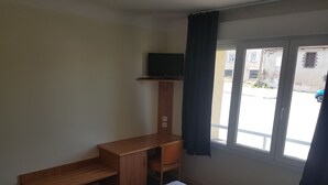 A hotel room with a bed, a wooden desk, a television on a shelf, and a window with curtains.