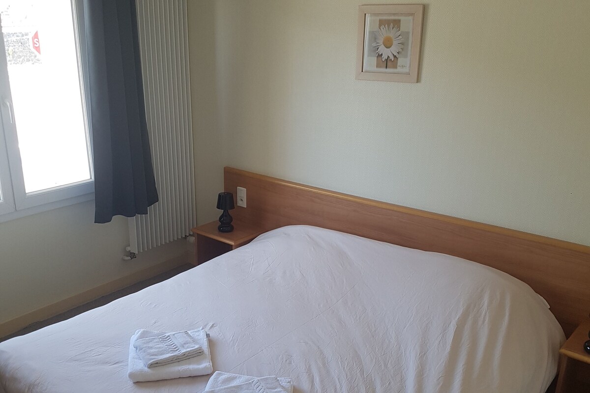 Een hotelkamer met een groot bed, een nachtkastje met een lamp, een raam met gordijnen en een lijst met een afbeelding aan de muur.
