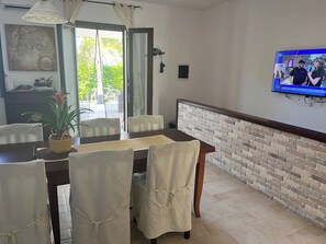Un coin repas agrémenté d’un mur en briques apparentes, d’un téléviseur fixé au-dessus d’une table et offrant une vue sur l’extérieur grâce à une porte vitrée.