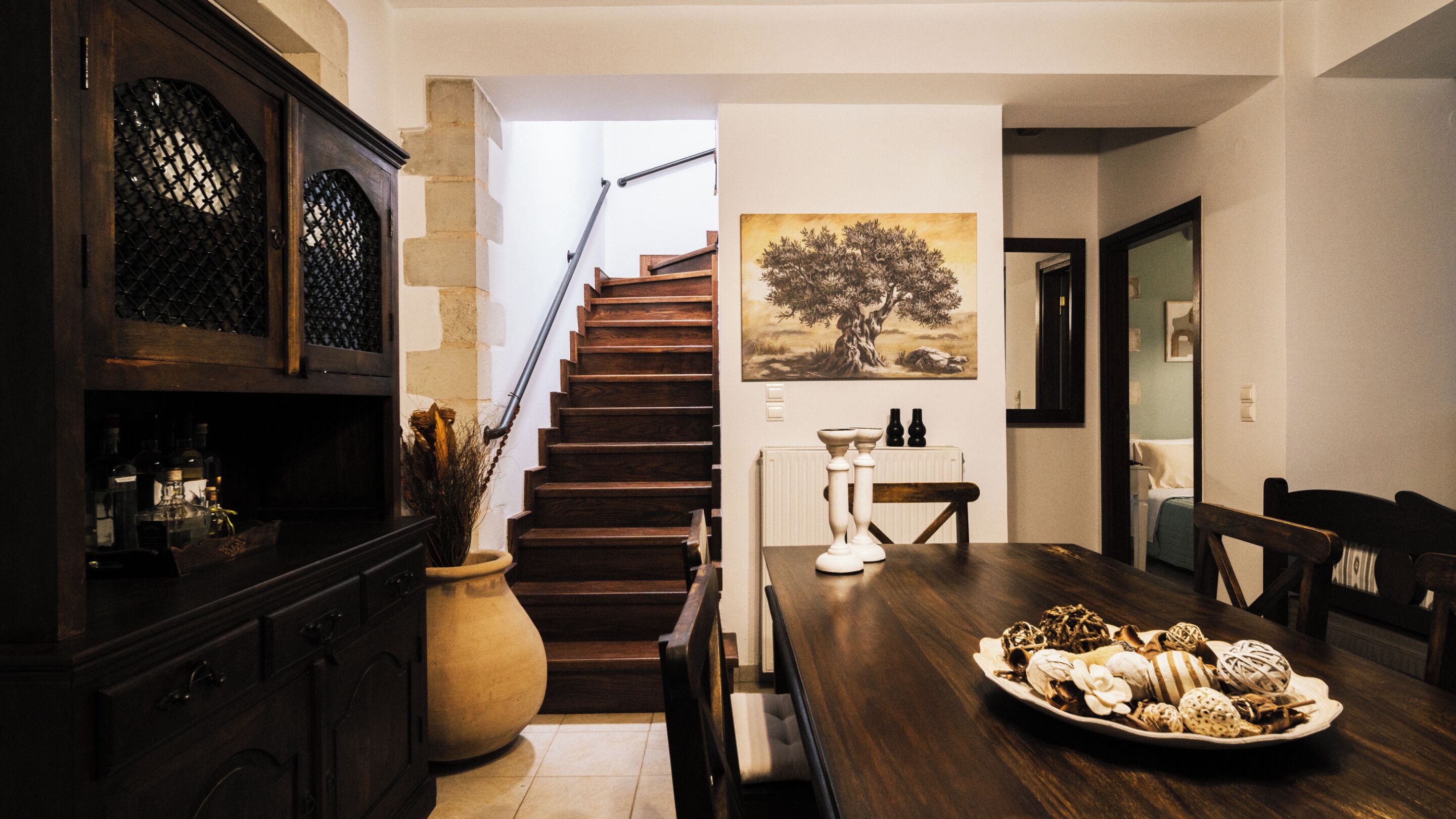 Uma sala de jantar com uma mesa de madeira posta para uma refeição, uma escada que leva a outro cômodo e um quadro de uma árvore na parede.