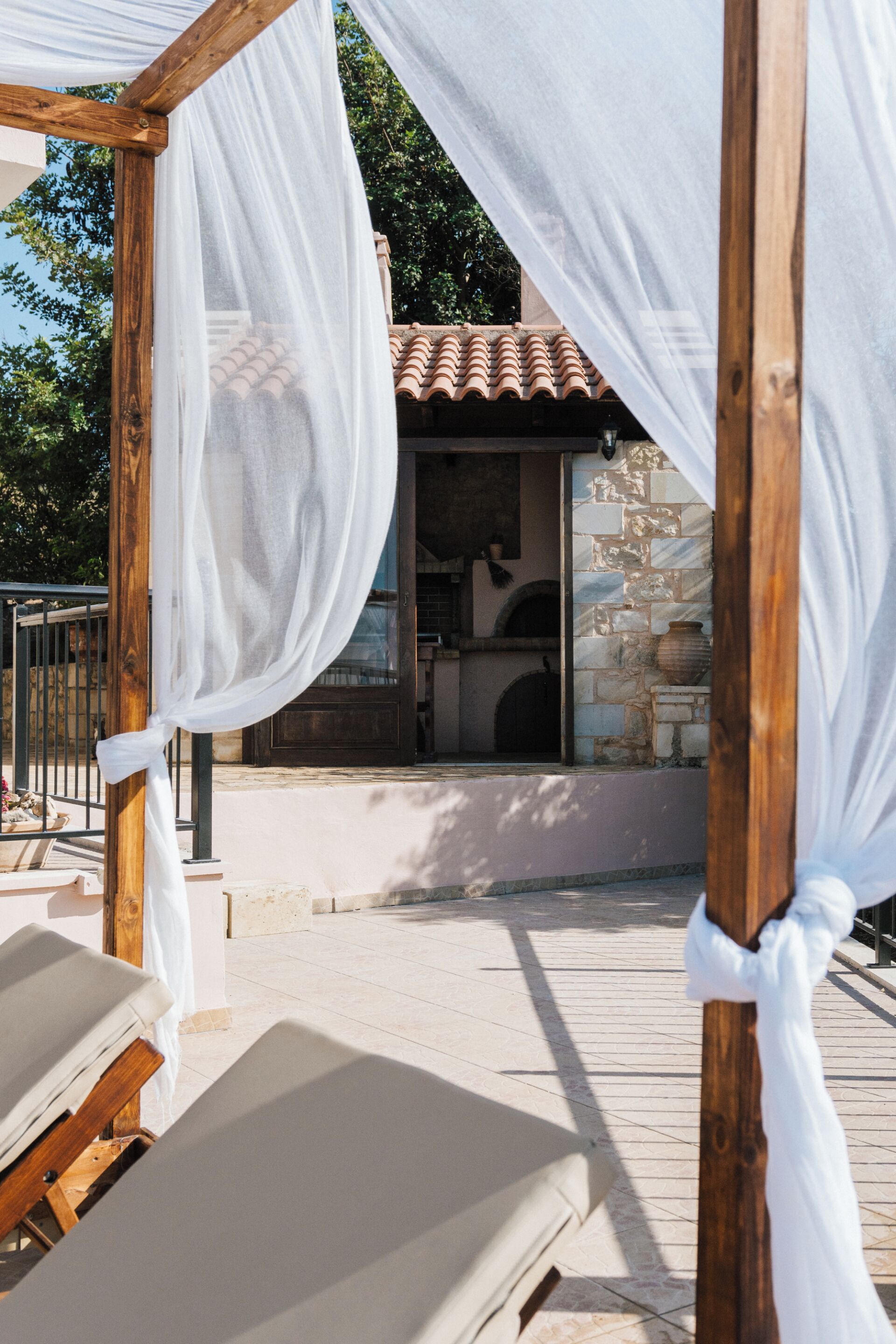 Una pérgola de madera con cortinas blancas, un edificio de piedra con techo de tejas y un suelo adoquinado.