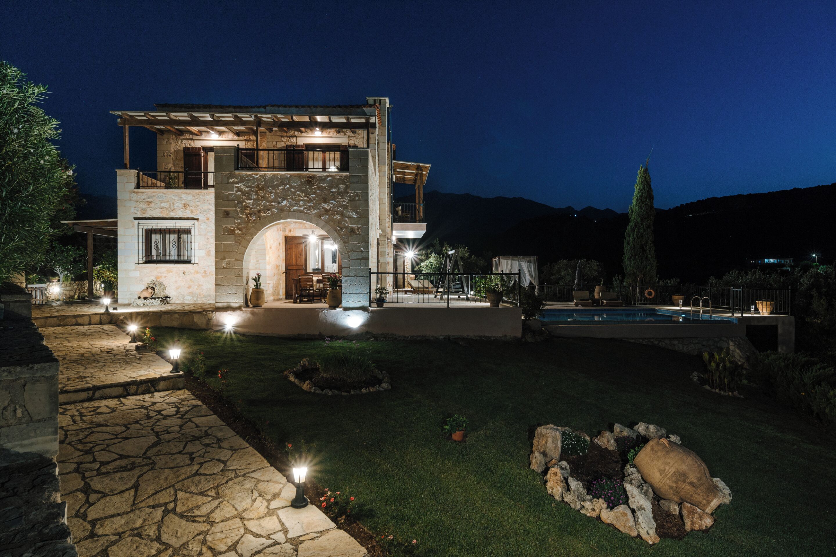Una casa de piedra de dos pisos con un arco de entrada de piedra, una piscina y un jardín bien iluminado.