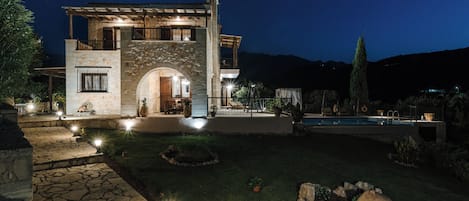 Una casa de piedra de dos pisos con un arco de entrada de piedra, una piscina y un jardín bien iluminado.