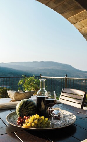 Uma mesa com uma garrafa, copos, uvas e melancia em um terraço com vista para as montanhas.
