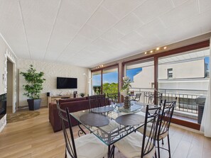 A spacious living area with a glass dining table, comfortable seating, a flat-screen TV, and a large window offering a view of the outdoors. - Seaside getaway for 2 with balcony (Les Sables-d´Olonne)