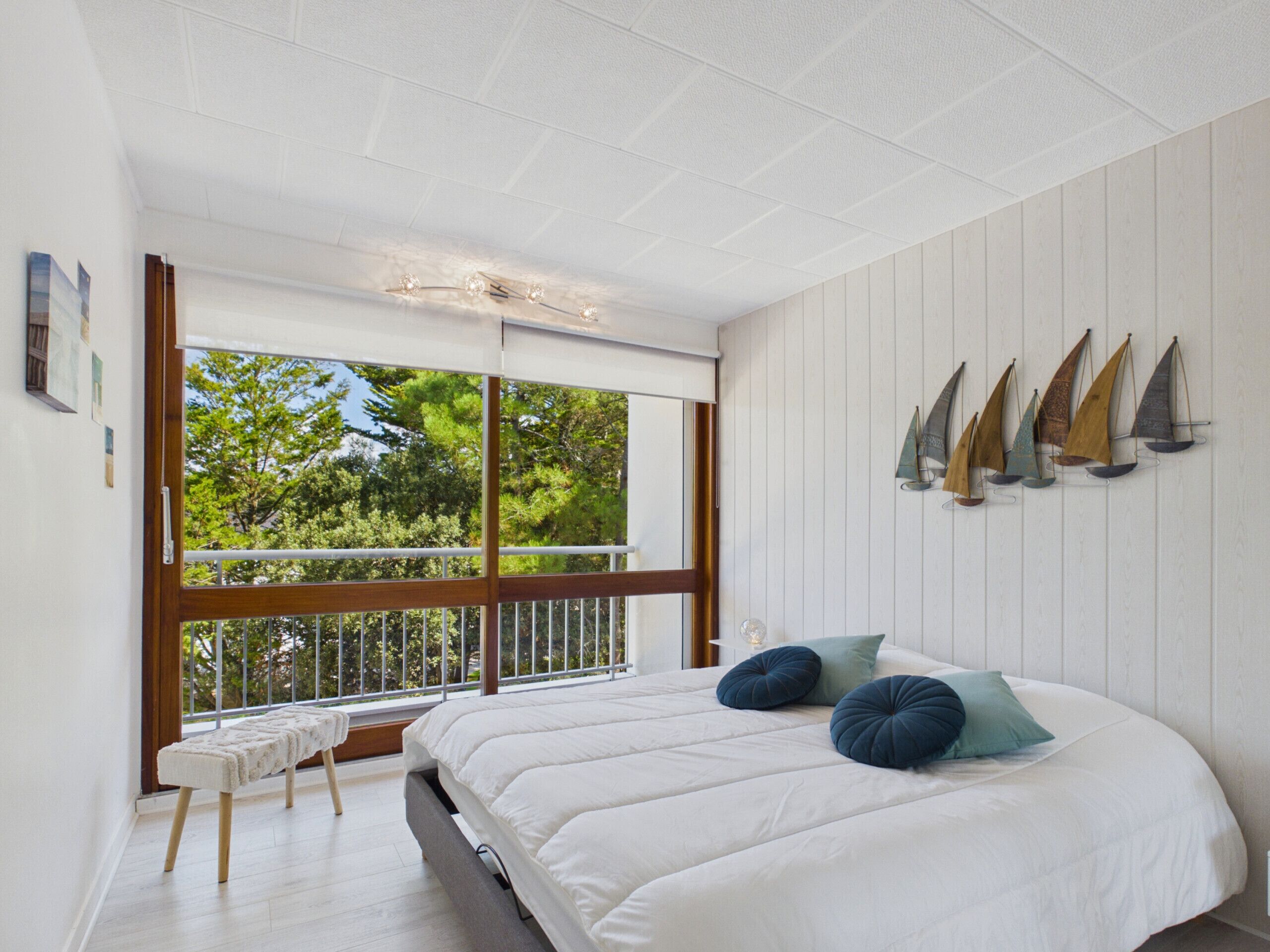 A bedroom with a large window, a bed with white bedding, and a bench.