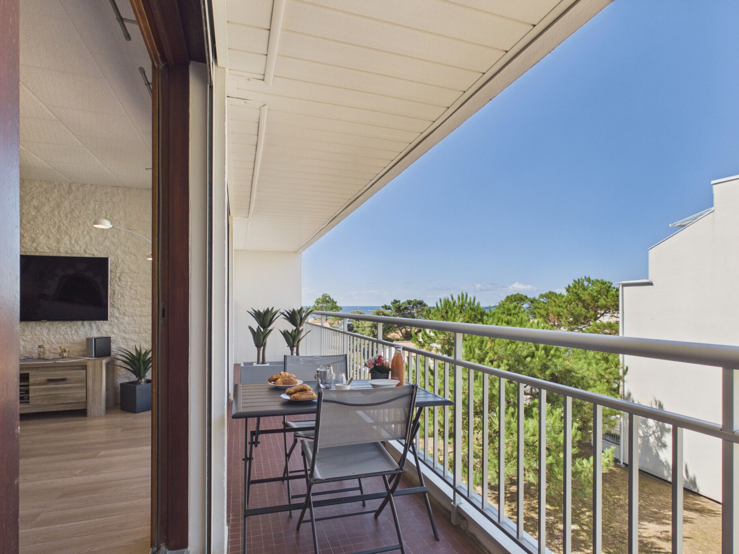 A balcony with a table and chairs, a TV mounted on the wall, and a view of the sea.