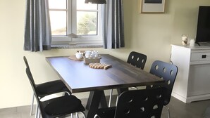 Uma sala de jantar com mesa, cadeiras, luminária, janela com cortinas, quadro na parede e televisão sobre um móvel.