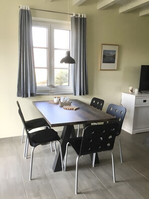 Ein Essbereich mit Tisch, Stühlen, einer Lampe, einem Fenster mit Vorhängen, einem Bild an der Wand und einem Fernseher auf einem Schrank.