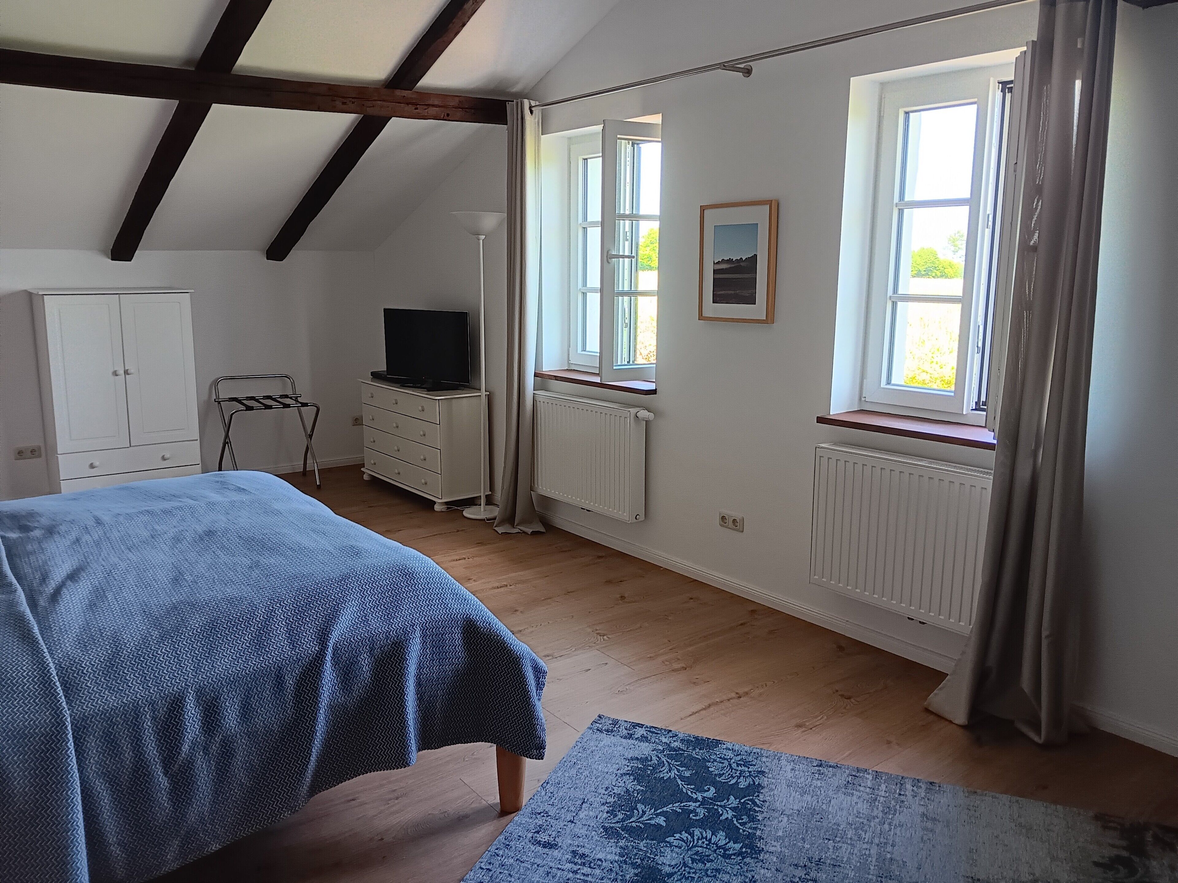 Ein Schlafzimmer mit einem Bett, einem Schminktisch, einem Fernseher und zwei mit Vorhängen versehenen Fenstern.