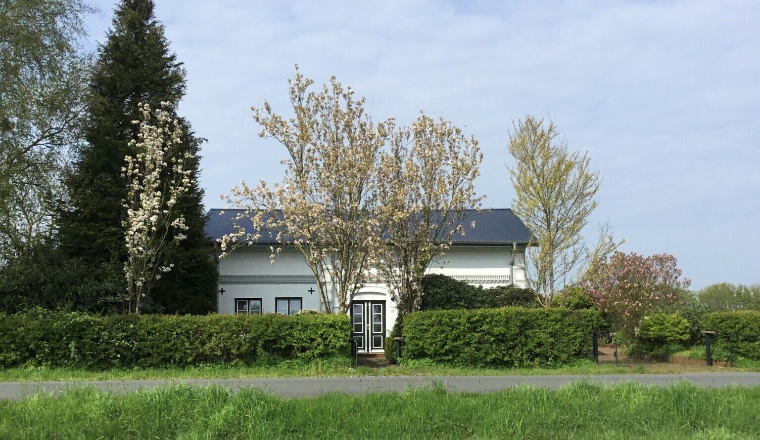 Ein zweigeschossiges Haus mit dunklem Dach, umgeben von gepflegten Hecken und blühenden Bäumen.