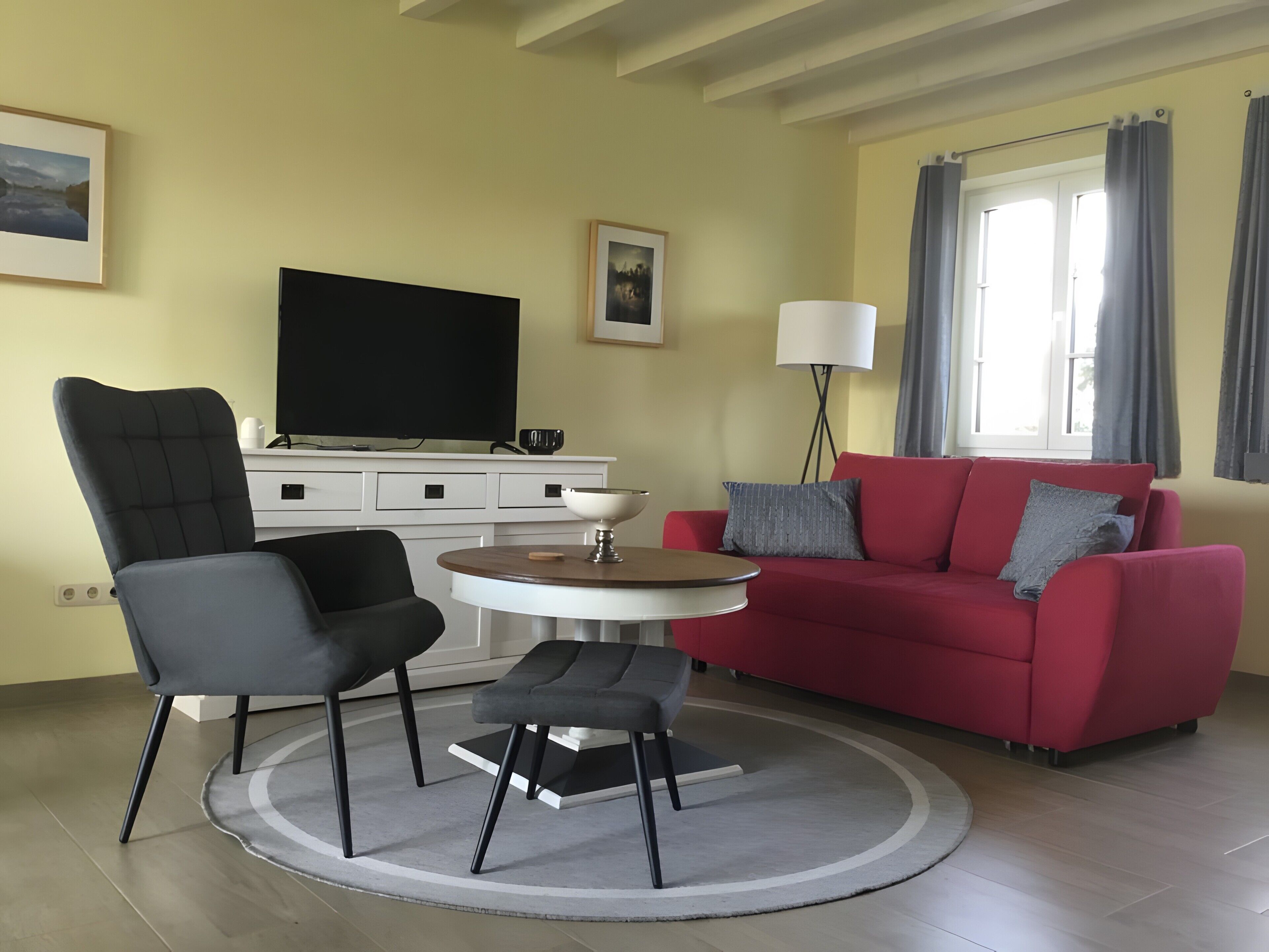 Ein Wohnzimmer mit Fernseher, einem roten Sofa, einem schwarzen Sessel, einem kleinen runden Tisch und einer Stehlampe.