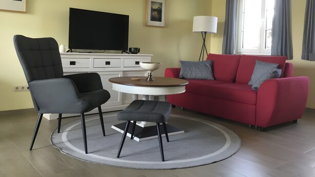 Uma sala de estar com televisão, um sofá vermelho, uma cadeira preta, uma mesa redonda pequena e um abajur.