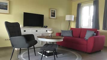 Ein Wohnzimmer mit Fernseher, einem roten Sofa, einem schwarzen Sessel, einem kleinen runden Tisch und einer Stehlampe.