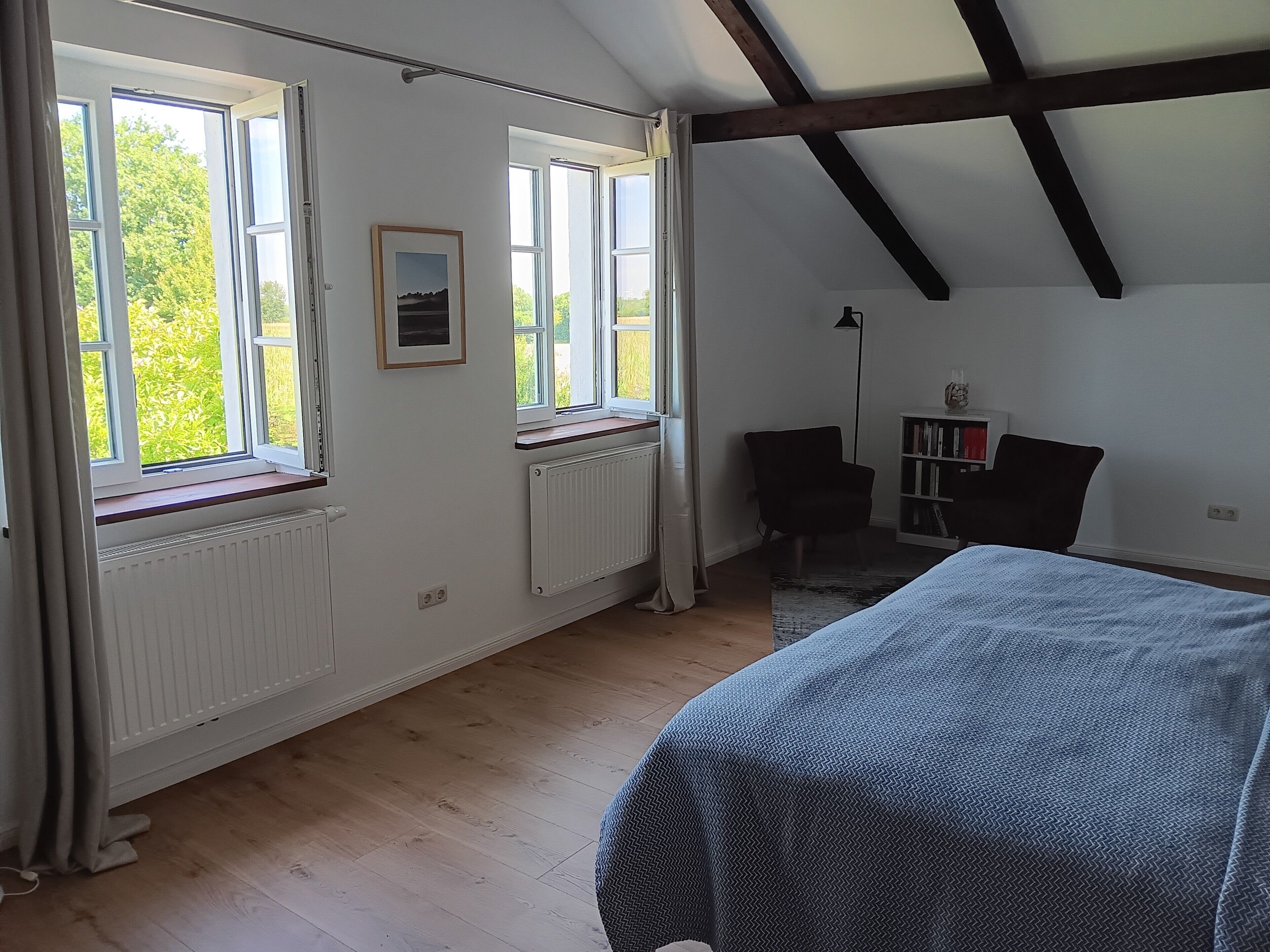 Ein Schlafzimmer mit einem großen Bett, zwei Fenstern, einem Heizkörper, einem Bücherregal und einem Sessel.