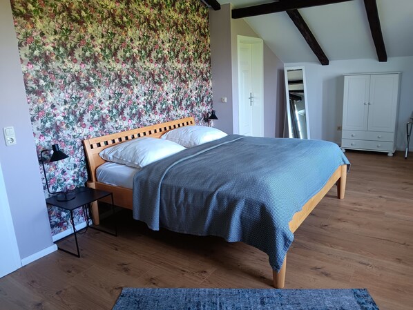 A bedroom with a wooden bed, a floral wallpaper, a white wardrobe, and a wooden floor.