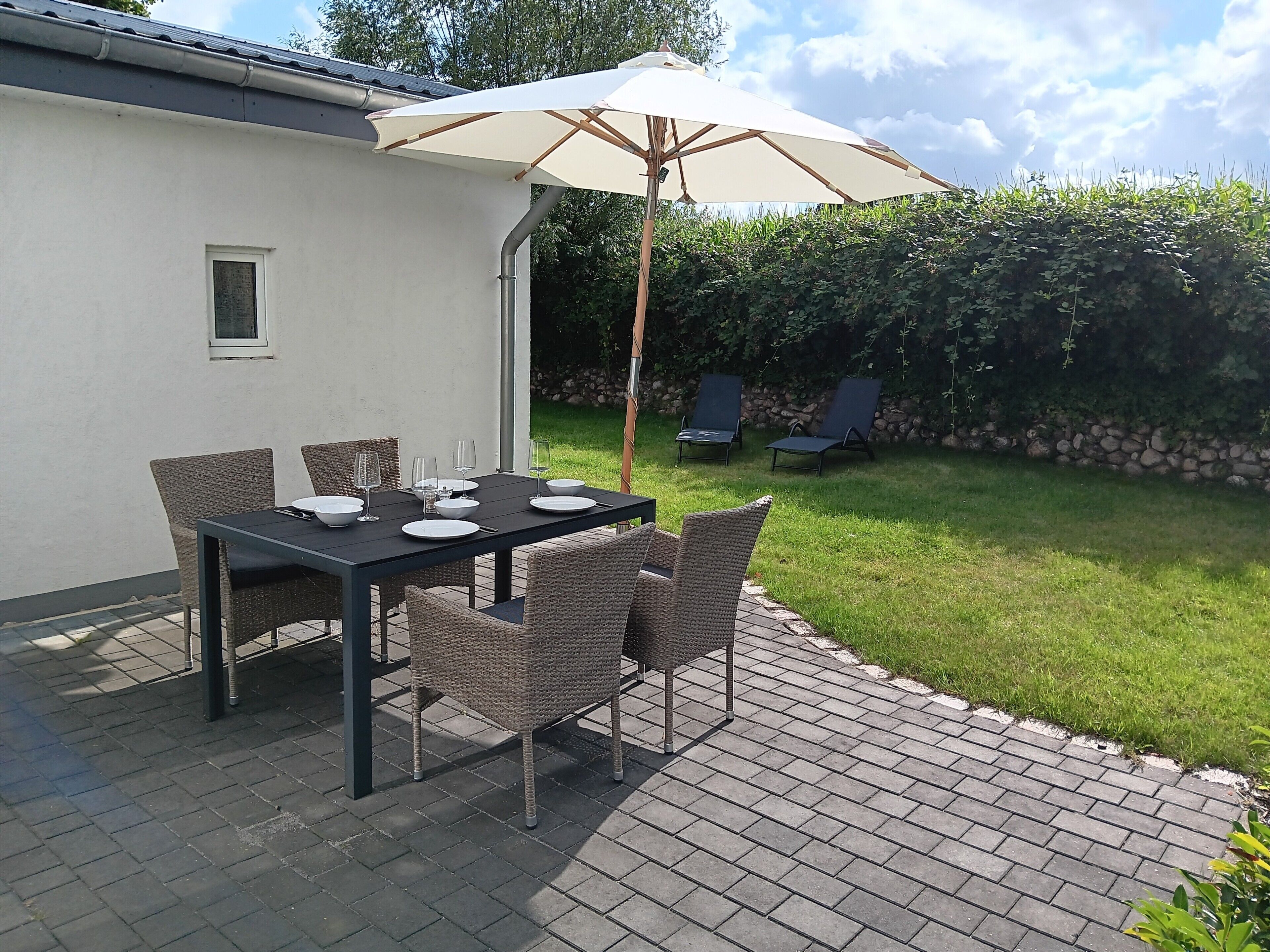 Eine Terrasse im Freien mit einem Esstisch, umgeben von Korbstühlen und einem Sonnenschirm, der Schatten spendet.