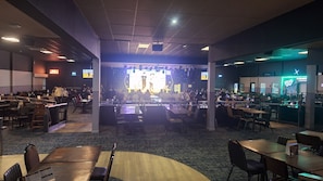 A spacious indoor dining area with multiple tables and chairs, a stage for performances, and a counter with a menu display. - Mkrj Premium - Parkdean Camber Sands - (Rye)