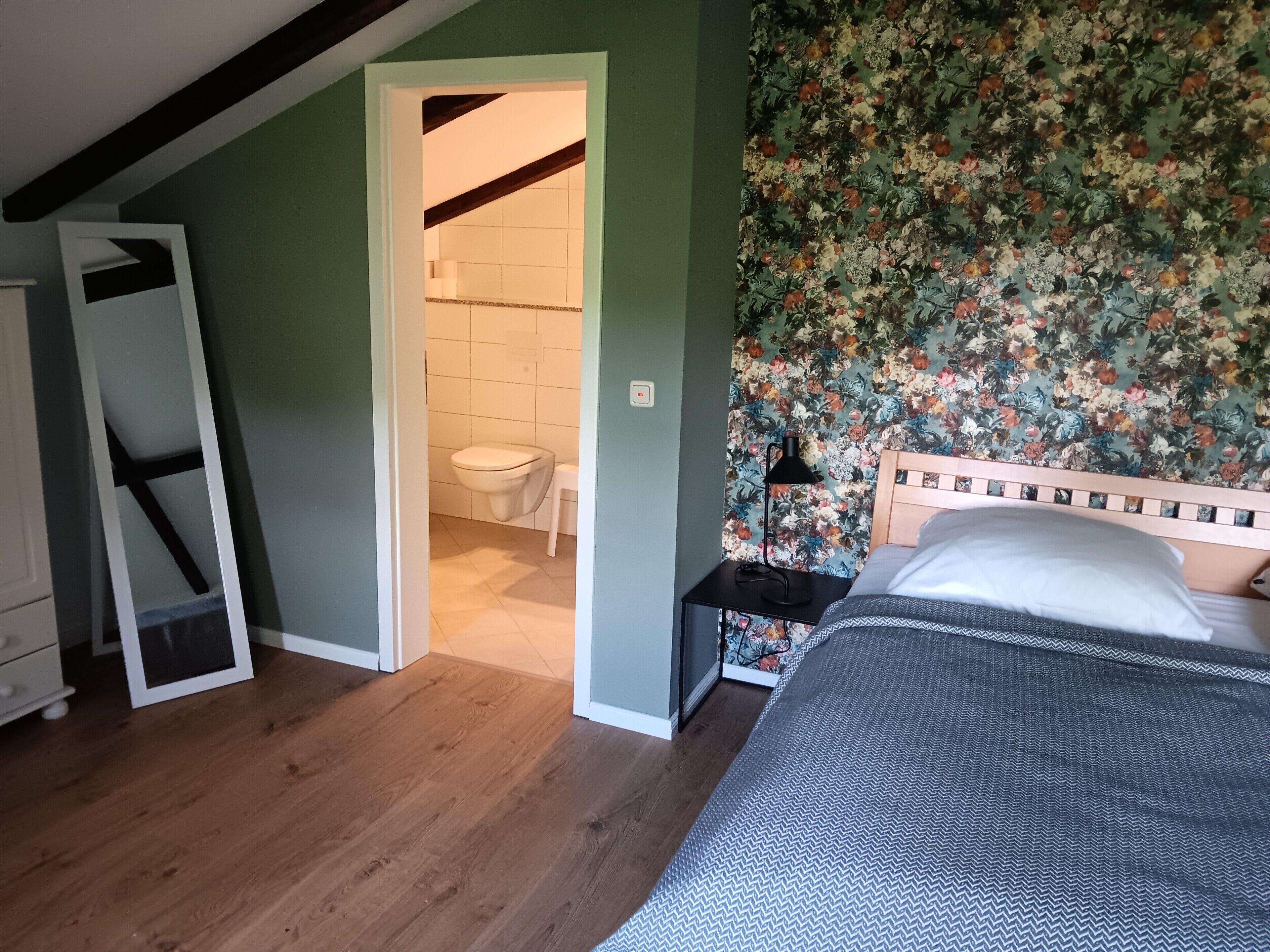 Ein Schlafzimmer mit einem Bett, einem Nachttisch, Holzboden, einer Tür zu einem Badezimmer und floralem Wandpapier.