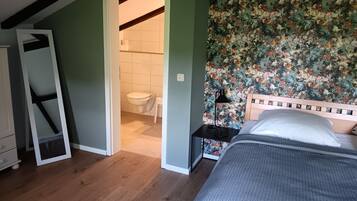 Ein Schlafzimmer mit einem Bett, einem Nachttisch, Holzboden, einer Tür zu einem Badezimmer und floralem Wandpapier.