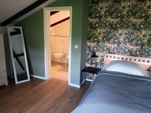 Ein Schlafzimmer mit einem Bett, einem Nachttisch, Holzboden, einer Tür zu einem Badezimmer und floralem Wandpapier.