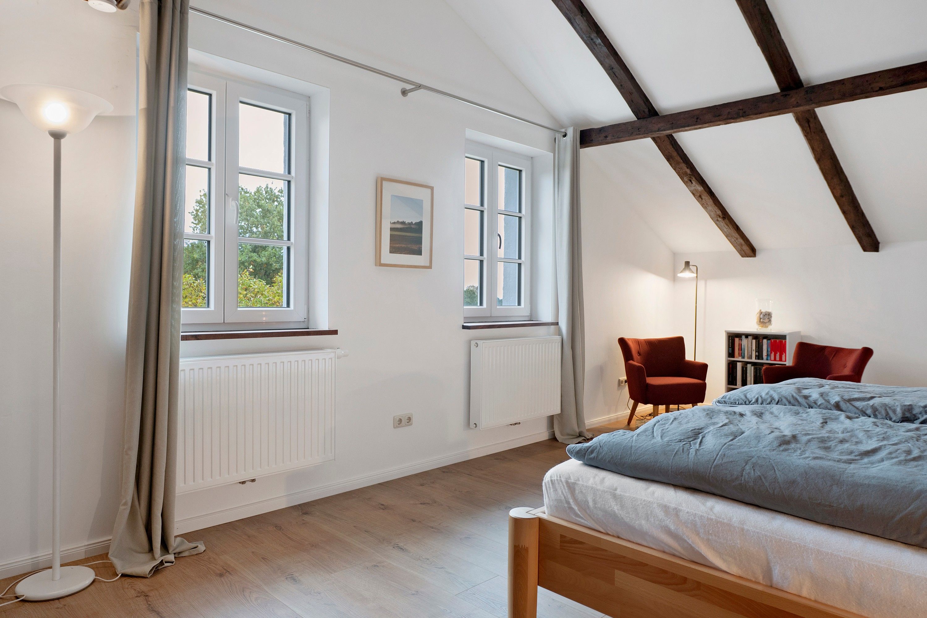 Ein Schlafzimmer mit einem Bett, zwei Stühlen, einer Stehlampe, einem Heizkörper und zwei Fenstern.