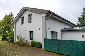 Un bâtiment blanc avec un toit à pignon et de nombreuses fenêtres, entouré d’une clôture verte et de quelques plantes.