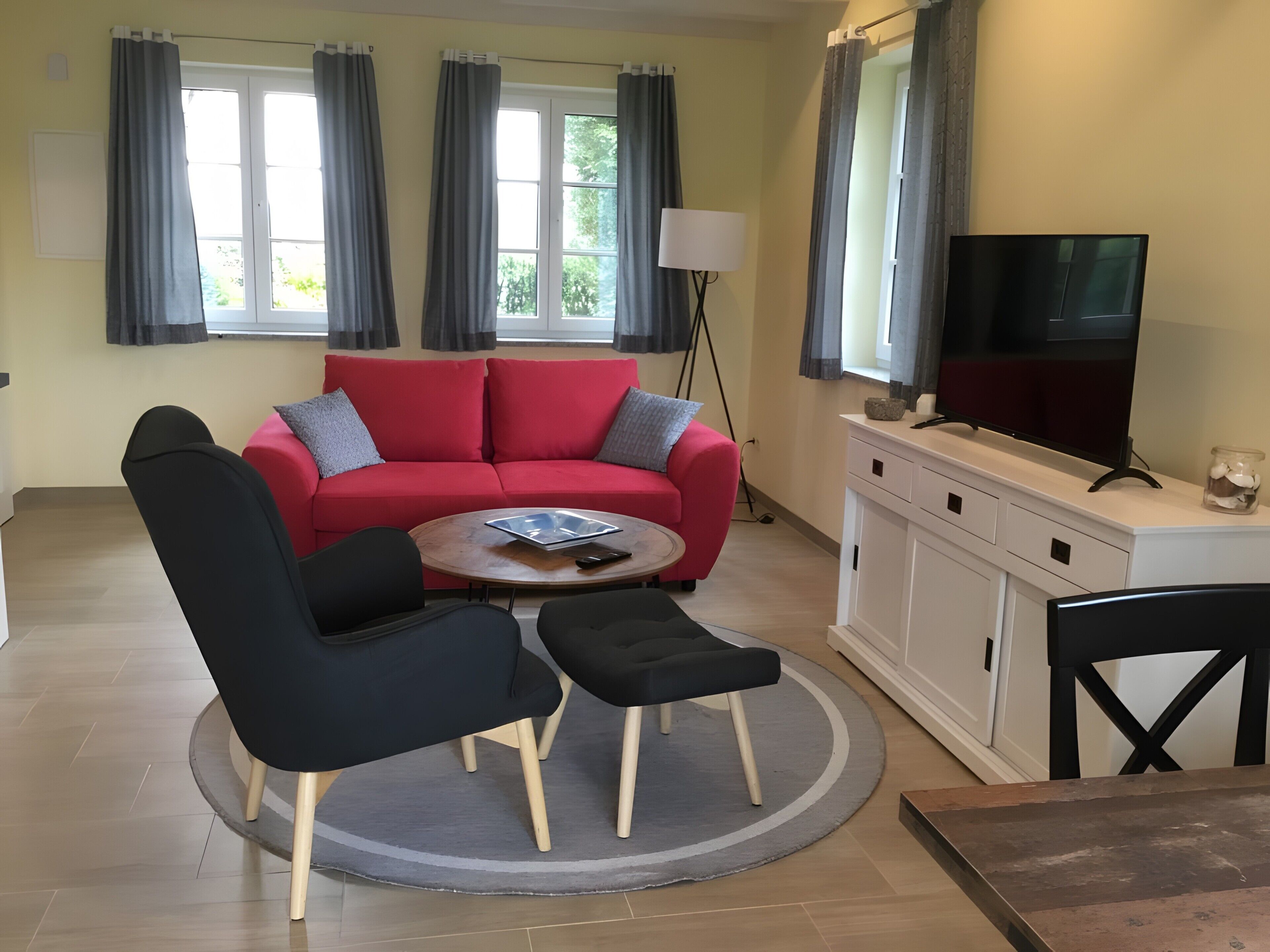 Ein Wohnzimmer mit einem roten Sofa, zwei schwarzen Sesseln, einem Couchtisch, einem Fernseher auf einer weißen Kommode und einer Lampe.