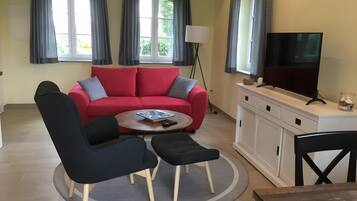 Ein Wohnzimmer mit einem roten Sofa, zwei schwarzen Sesseln, einem Couchtisch, einem Fernseher auf einer weißen Kommode und einer Lampe.