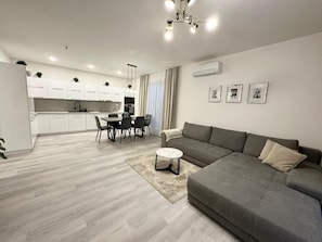 A modern living room with a grey sofa, a dining area with a table and chairs, and a kitchen in the background.