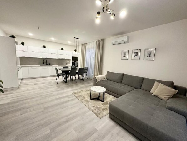 Un salon moderne avec un canapé gris, un coin repas avec une table et des chaises, et une cuisine en arrière-plan.