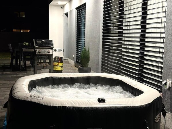 A hot tub with a pump, a patio area with chairs, and a building with large windows in the background.