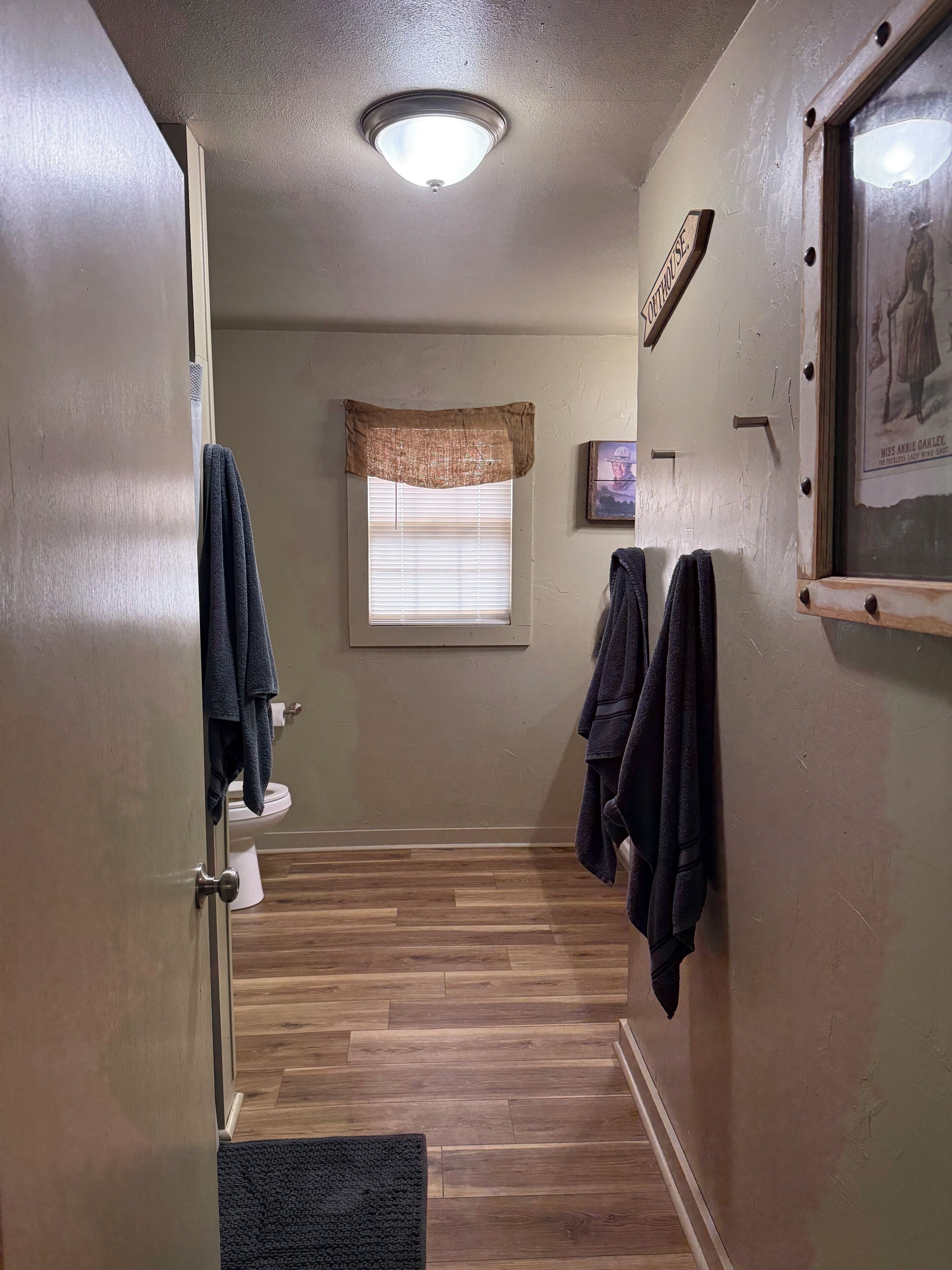 A narrow bathroom with a wooden floor, a toilet, a towel rack, and a window with blinds.