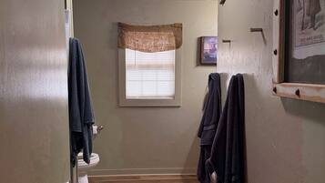 A narrow bathroom with a wooden floor, a toilet, a towel rack, and a window with blinds.