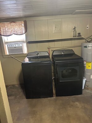 Une buanderie équipée de deux machines à laver et deux sèche-linge superposés, d’une fenêtre avec des stores, d’un radiateur et d’un chauffe-eau.