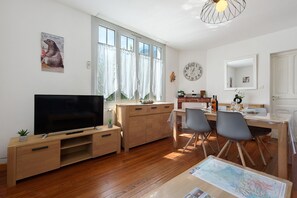 A modern living room with a flat-screen TV, wooden furniture, a dining area with chairs, a table, and a large window with curtains. - “Gîte de l'isard” Apartment: Mountain View, Private Garden, Wi-Fi (Pierrefitte-Nestalas)