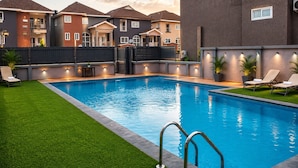 Piscina com um deck ao redor, cadeiras de descanso e vista para edifícios residenciais.