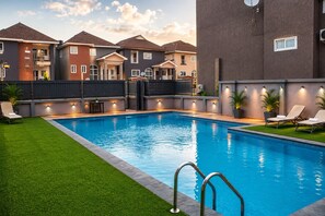 Une piscine entourée d’une terrasse, de chaises longues et offrant une vue sur des immeubles résidentiels.