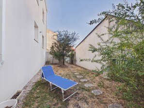 Ein schmaler Außenkorridor mit einer blauen Liege, einem Baum und einem Gebäude mit weißer Fassade.