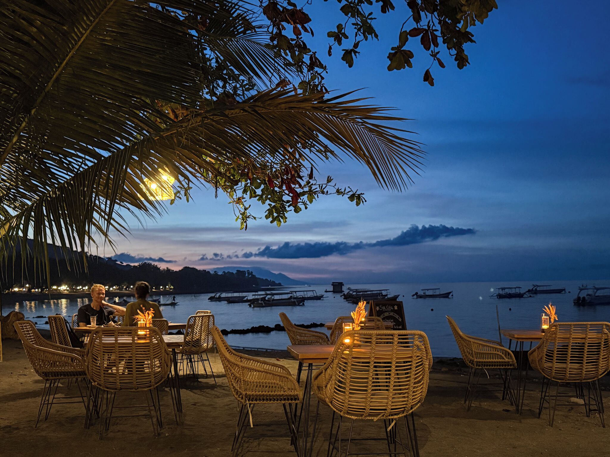 Un espace extérieur aménagé avec des chaises et des tables en osier, offrant une vue sur une mer calme, des bateaux et un paysage montagneux en arrière-plan.