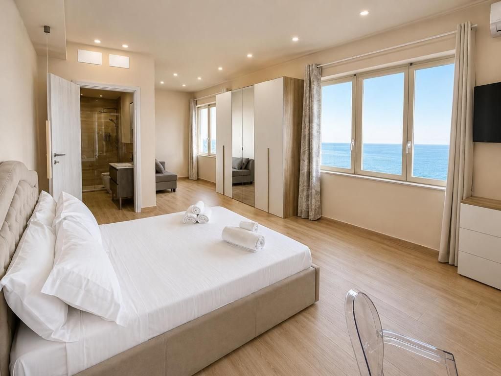 A bedroom with a large bed, a walk-in closet, a bathroom, and a view of the sea.