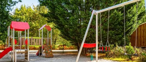 Un parque infantil con columpios y toboganes, rodeado de árboles y una estructura de madera.