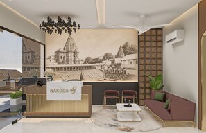 A reception area with a reception desk, two chairs, a sofa, a chandelier, and a wall with a cityscape mural. - Hotel Parshwa (Ujjain)